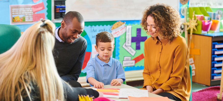 A young child sits at a classroom desk with a supportive adult to his left leaning in to look at his work and another adult sat admiring his work on the right.